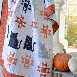 May include: A white quilt with a black and orange star pattern and black cat silhouettes. The quilt is draped over a white railing with two orange pumpkins in the foreground.
