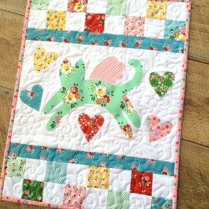 May include: A white quilted wall hanging with a green cat applique. The cat is surrounded by pink, red, and yellow hearts. The background is a patchwork of squares in various colors and patterns.