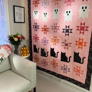 May include: A pink and black quilt with a Halloween theme. The quilt features a repeating pattern of stars, ghosts, and black cats. The quilt is displayed on a wall in a room with a couch and a small table.