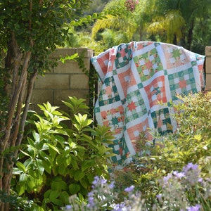 May include: A patchwork quilt with a green, white, and pink color scheme. The quilt is draped over a brick wall and surrounded by green foliage.