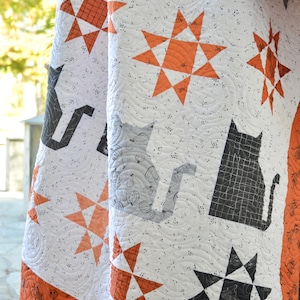 May include: A white quilt with a black and orange star pattern. The quilt has a black cat and pumpkin design on one side and a black cat silhouette on the other side.