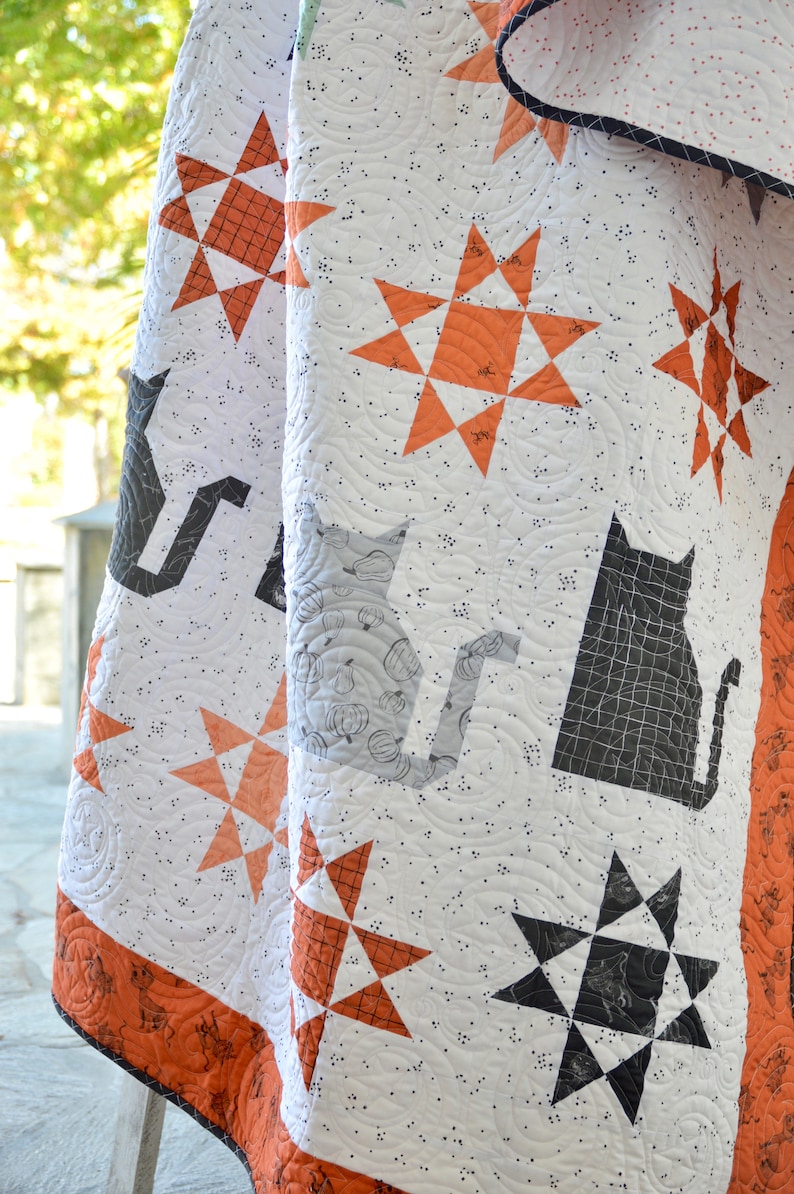 May include: A white quilt with a black and orange star pattern. The quilt has a black cat and pumpkin design on one side and a black cat silhouette on the other side.