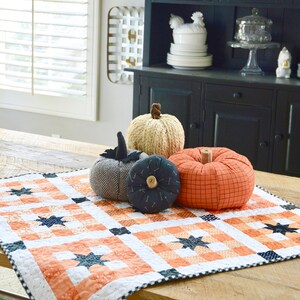 May include: A black and white checkered table runner with orange stars on a wooden table. There are four fabric pumpkins on the table runner, one is beige, one is orange, and two are black.