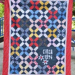 May include: A red, white, and blue patchwork quilt with a black background. The quilt is made of squares with different patterns, including polka dots, gingham, and floral prints. The quilt has a red border and a white inscription that reads "CIRCA 1974".