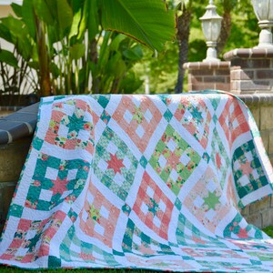 May include: A pink, green, and white quilt with a diamond pattern. The quilt is folded and lying on a green lawn.