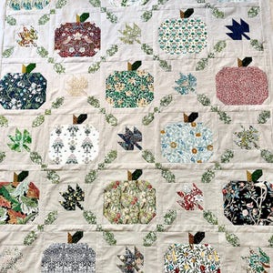 Pode incluir: Uma colcha com um design de patchwork que apresenta vários padrões florais e folhas verdes. A colcha é feita de quadrados com um motivo central de uma maçã, rodeada por uma borda de folhas. A colcha é de cor bege com uma textura subtil.