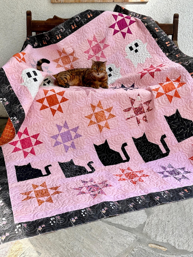 May include: A pink and black quilt with a cat sitting on it. The quilt features a variety of patterns, including stars, ghosts, and cats. The quilt is made with a variety of fabrics, including cotton and flannel.