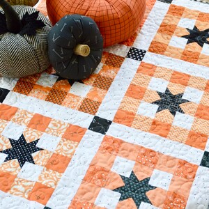 May include: A white and orange patchwork quilt with black stars. The quilt is decorated with pumpkins and is sitting on a wooden table.