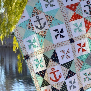 May include: A patchwork quilt with a nautical theme. The quilt is made of various fabrics in shades of blue, red, white, and grey. The quilt features anchors, pinwheels, and stars. The quilt is hanging from a tree branch over a body of water.