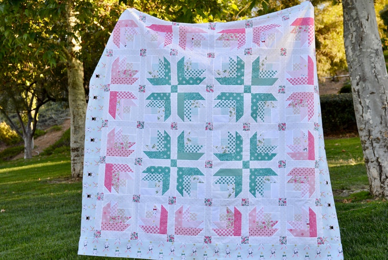 K&ouml;nnte beinhalten: Eine wei&szlig;e Steppdecke mit einem rosa-gr&uuml;nen Blumenmuster. Die Steppdecke besteht aus Quadraten mit einem sich wiederholenden Muster aus vierzackigen Sternen. Die Steppdecke h&auml;ngt an einer W&auml;scheleine in einem grasbewachsenen Bereich.