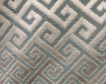 May include: Close-up of a fabric with a repeating geometric pattern. The design features a series of interlocking, stylized Greek key motifs in shades of light blue, gray, and off-white. The fabric has a textured appearance.