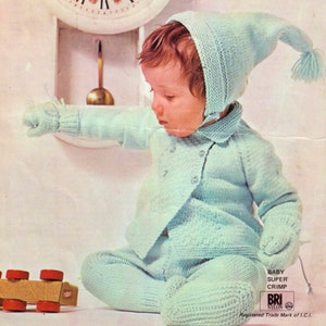May include: A baby wearing a light blue knitted outfit, including a hat, sweater, trousers, and mittens. The outfit is made with a textured stitch and has a button fastening on the sweater. The baby is sitting on a white surface and is reaching for a wooden toy.