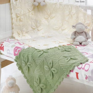 May include: Two knitted blankets in cream and sage green, each featuring a square-based floral motif with raised petals and a scalloped edge. The blankets are displayed on a surface with a printed alphabet pattern, accompanied by two plush toys.