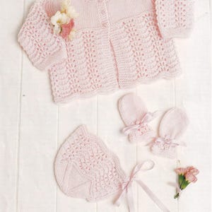 Könnte beinhalten: Ein rosa gestricktes Baby-Set bestehend aus einer Strickjacke, einer Mütze und Fäustlingen. Das Set hat ein zartes Spitzenmuster.