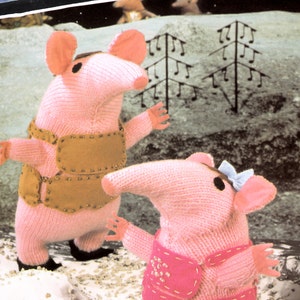 Puede incluir: Dos personajes de juguete de punto rosa del programa de televisión infantil "Clangers". Los personajes llevan chalecos marrones y rosas y tienen narices largas. Los personajes están de pie sobre una superficie blanca con pequeñas piedras.