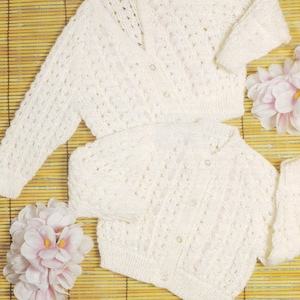 以下が含まれることがあります： ボタン留めの白いクロシェ編みのカーディガン2枚。カーディガンは、ピンクの花が置かれた竹製の編まれた表面に置かれています。