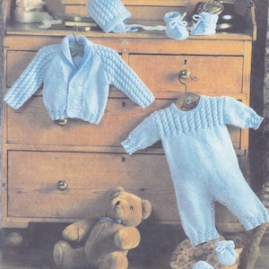 May include: A blue knitted baby outfit set including a hat, cardigan, and one-piece sleeper with booties. The set is displayed on a wooden dresser with a teddy bear.