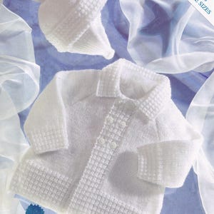 May include: A white knitted baby outfit, including a button-up cardigan and a matching hat with a pom-pom. The outfit is displayed on a blue background with the text "INCLUDES PREMATURE SIZES". The cardigan has a textured collar and cuffs.