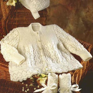 May include: A white knitted baby outfit set including a cardigan, bonnet, and booties. The cardigan has a button closure and a delicate lace pattern. The bonnet has a scalloped edge. The booties have a bow detail.