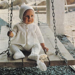 May include: A baby wearing a white knitted outfit, including a bonnet, jacket, pants, and booties, sits on a wooden swing. The outfit has a textured pattern and button details. The swing is suspended by metal chains.