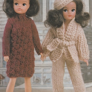 May include: Two dolls wearing knitted outfits. One doll is in a burgundy dress, and the other is in a beige pantsuit with a matching beret. The dolls are holding hands, set against a backdrop of a cityscape.