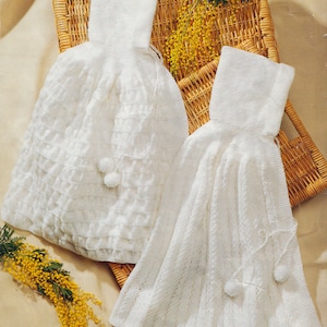 Op de afbeelding: Twee witte gebreide kledingstukken met een hoge hals en een uitlopend model. Het ene kledingstuk heeft een gestructureerd ruitpatroon, terwijl het andere verticale ribbeldetails heeft. Beide items hebben dunne witte koordjes met pompons.
