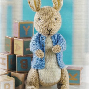 May include: A knitted bunny toy with long ears, wearing a blue jacket over a white body. The bunny is standing in front of a stack of wooden alphabet blocks with various letters and colours.