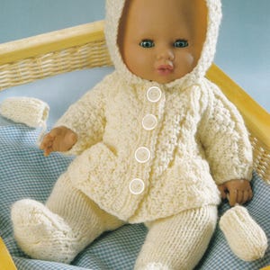 May include: A doll dressed in a cream-coloured knitted outfit, including a hooded jacket with four buttons, trousers, and mittens. The doll is in a wooden basket with a blue and white checked liner.
