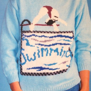 May include: A light blue knitted sweater with a graphic of a swimmer jumping into a pool. The graphic includes the word "Swimwear" in blue lettering.