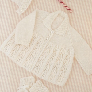 May include: A cream-coloured baby clothing set, comprising a long-sleeved sweater with a collar and button closure, a bonnet, and a pair of booties with ribbon ties. The sweater has a textured, openwork design. The set is displayed on a striped background.