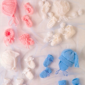 Può includere: Una collezione di vestiti per neonati fatti a mano in vari colori. L'immagine presenta cappellini, guanti e scarpine lavorate a maglia in rosa, bianco e blu. Alcune scarpine hanno fiocchi. Gli articoli sono disposti su uno sfondo chiaro.