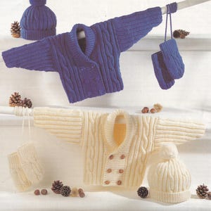 May include: A display of knitted children's clothing in blue and cream. The blue set includes a cable-knit cardigan, hat, and mittens. The cream set features a cardigan, hat, and mittens. Pinecones and nuts are scattered around the garments.