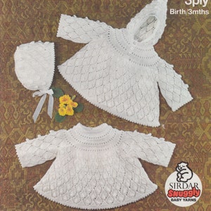 Puede incluir: Un cárdigan blanco de punto para bebé y un gorro a juego con un delicado patrón de encaje. El cárdigan tiene capucha y cierre de botones. El gorro tiene la parte superior fruncida y se ata debajo de la barbilla. La lana está etiquetada como "Sirdar Snuggly Baby Yarns".