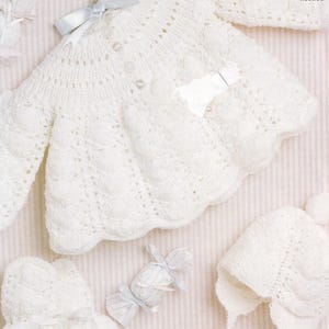 Puede incluir: Conjunto de ropa de bebé tejido a mano en color blanco, que incluye un cárdigan, un gorro y patucos. El cárdigan tiene un borde festoneado y cierre de botones. Se muestra sobre un fondo a rayas con una cinta azul claro.