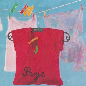 May include: A red garment with the word "Pegs" in cursive script is hanging on a black hanger. Clothes are hanging on a clothesline with colorful clothespins. The background is a light blue color.