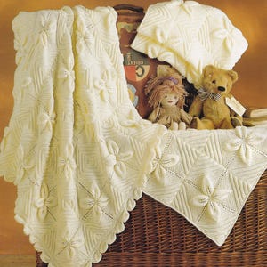 May include: A white knitted baby blanket with a floral pattern. The blanket is folded and draped over a wicker basket. There are two stuffed toys, a teddy bear and a doll, sitting on the blanket.