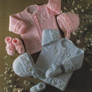 May include: A collection of knitted baby clothing in soft pink and light blue. The set includes sweaters, bonnets, and booties, all adorned with delicate floral embroidery and ribbon accents. The garments are arranged on a textured surface.