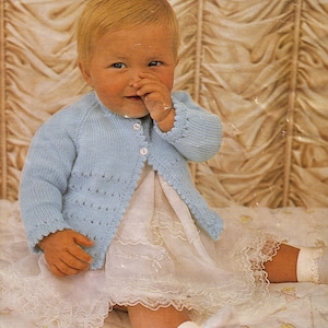 Puede incluir: Un cárdigan de bebé de punto azul claro con cierre de botones. El cárdigan tiene un delicado diseño calado en la parte inferior y en los puños. El bebé lleva un vestido blanco con ribete de encaje.