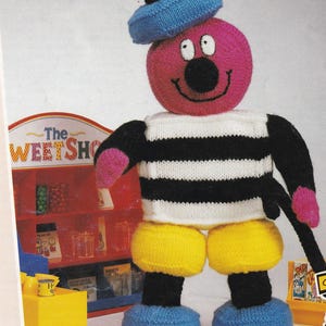 Può includere: Una bambola di maglia con una testa rosa, un naso nero e un cappello blu. La bambola ha una camicia a righe bianche e nere, pantaloncini gialli e scarpe blu. La bambola tiene in mano un bastone nero. Lo sfondo include un cartello con la scritta "The Sweet Shop".
