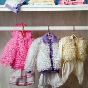 May include: Three knitted baby outfits hanging on a white rack. The outfits are a pink cardigan with a white skirt, a white cardigan with a purple button-up shirt and white shorts, and a yellow cardigan with a white shirt and pants. The outfits are all made with a fluffy, textured yarn.