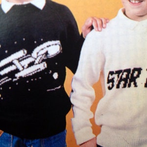 May include: Two children wearing knitted sweaters. The child on the left is wearing a black sweater with a white spaceship design. The child on the right is wearing a white sweater with the text "STAR TREK" in black.
