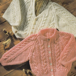 May include: Two knitted baby cardigans, one white and one pink, with a cable knit pattern. Both cardigans have button closures.