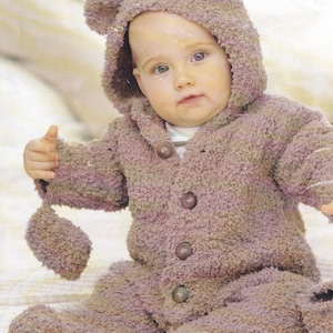 May include: A baby wearing a brown knitted bear costume with a hood and attached mittens. The costume has a button fastening down the front.