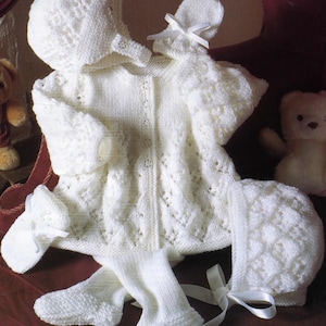 May include: A complete white knitted baby outfit, including a jacket, bonnet, booties, mittens, and leggings. The set features a delicate pattern and is tied with white ribbons. The ensemble is designed for a newborn.