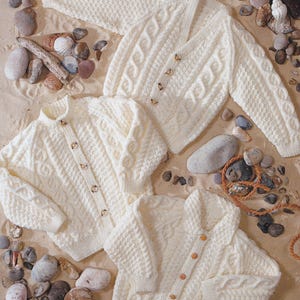 May include: Cream-colored cable-knit sweaters with wooden buttons are displayed on a sandy surface with pebbles and shells. The sweaters have various necklines and button arrangements, showcasing the intricate knit patterns. The scene evokes a cozy, handcrafted aesthetic.