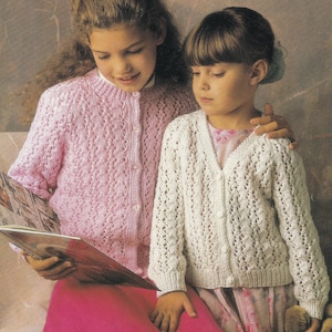May include: Two young girls wearing knitted cardigans. One girl wears a light pink cardigan with a button-up front, and the other wears a white cardigan. Both cardigans have a delicate, openwork design. The girls are looking at a book.