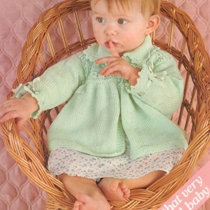 Puede incluir: Un bebé en una silla de mimbre con un cárdigan de punto verde claro y una falda a juego. El cárdigan tiene un cuello y puños con volantes y lazos pequeños. El texto "For that very special baby" es visible.