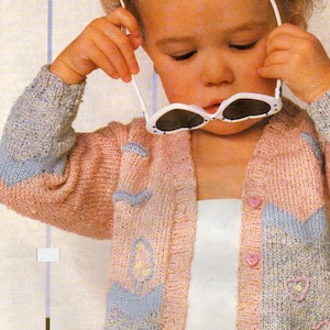 May include: A young child wearing a pink, blue, and grey knitted cardigan with a white button-up shirt underneath. The cardigan has a floral design and the child is wearing white sunglasses.