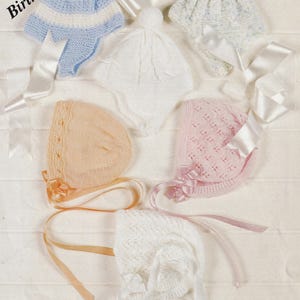 May include: Five knitted baby hats in different colors and styles, including white, pink, blue, and peach. The hats are displayed on a white background with satin ribbons.