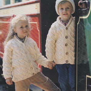 Puede incluir: Dos niños con cárdigans de punto de color crema con un patrón de rombos. La niña lleva pantalones marrones y el niño lleva pantalones vaqueros azules. Están de pie frente a una puerta verde y negra.